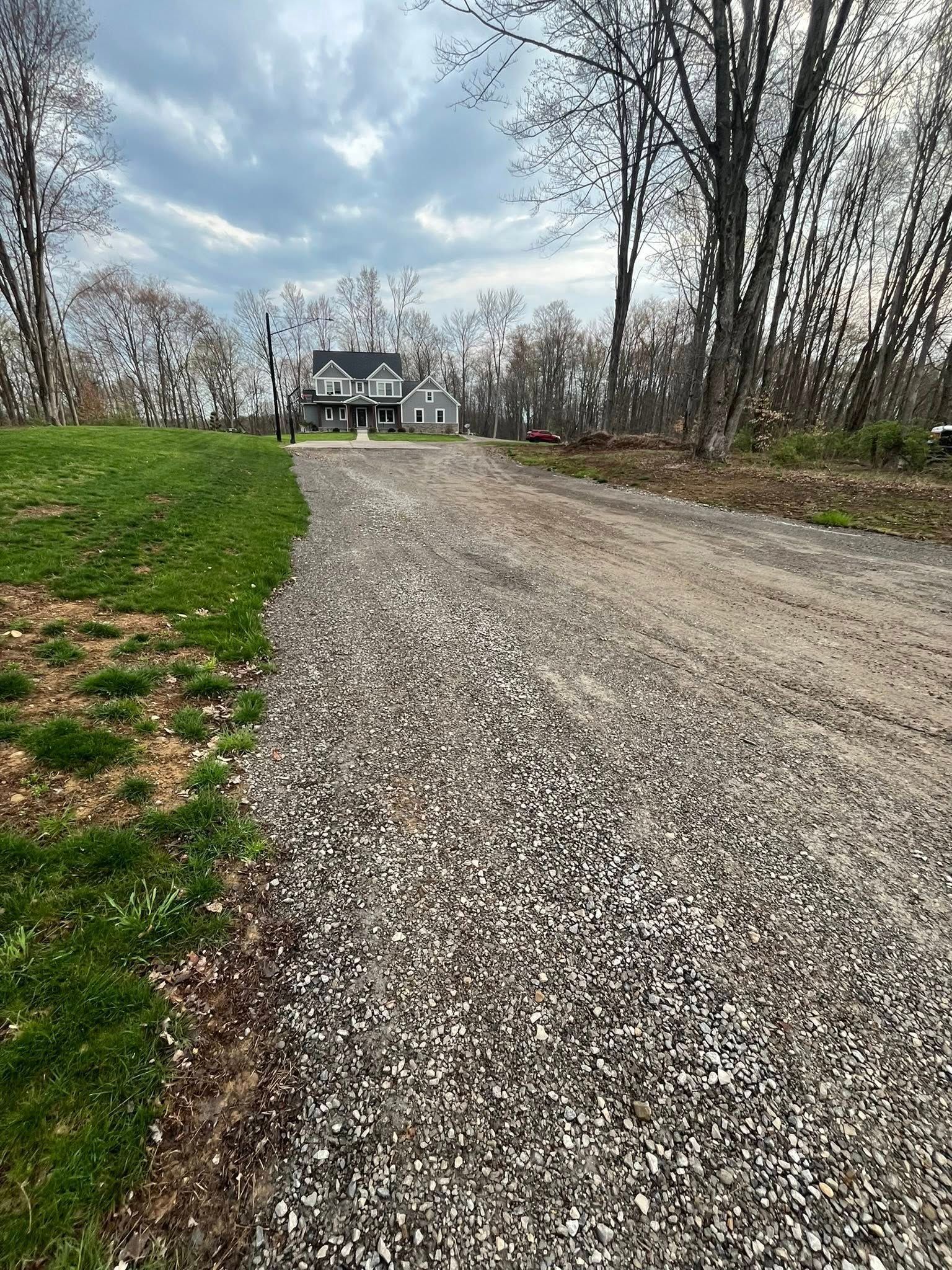 A gravel driveway leads toward a two-story house surrounded by trees under a cloudy sky.