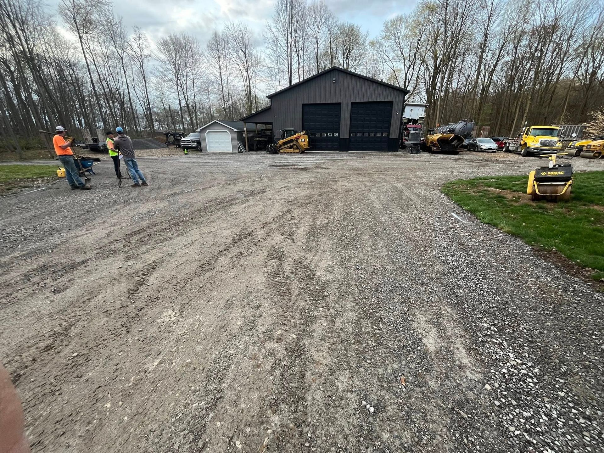 A gravel lot with three people standing on the left, a dark warehouse in the distance, and construction equipment nearby.