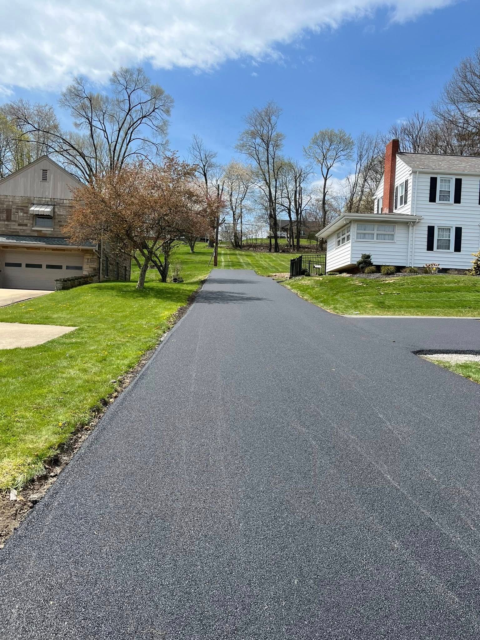 A newly paved, dark asphalt driveway leads toward a white house and a separate garage on a sunny, grassy lawn.