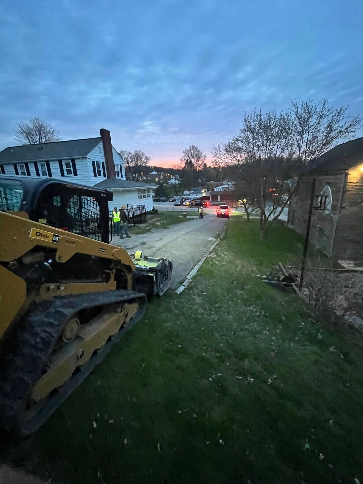 A yellow skid steer loader sits on a road at dusk, near a house, with a person wearing a high-visibility vest nearby.