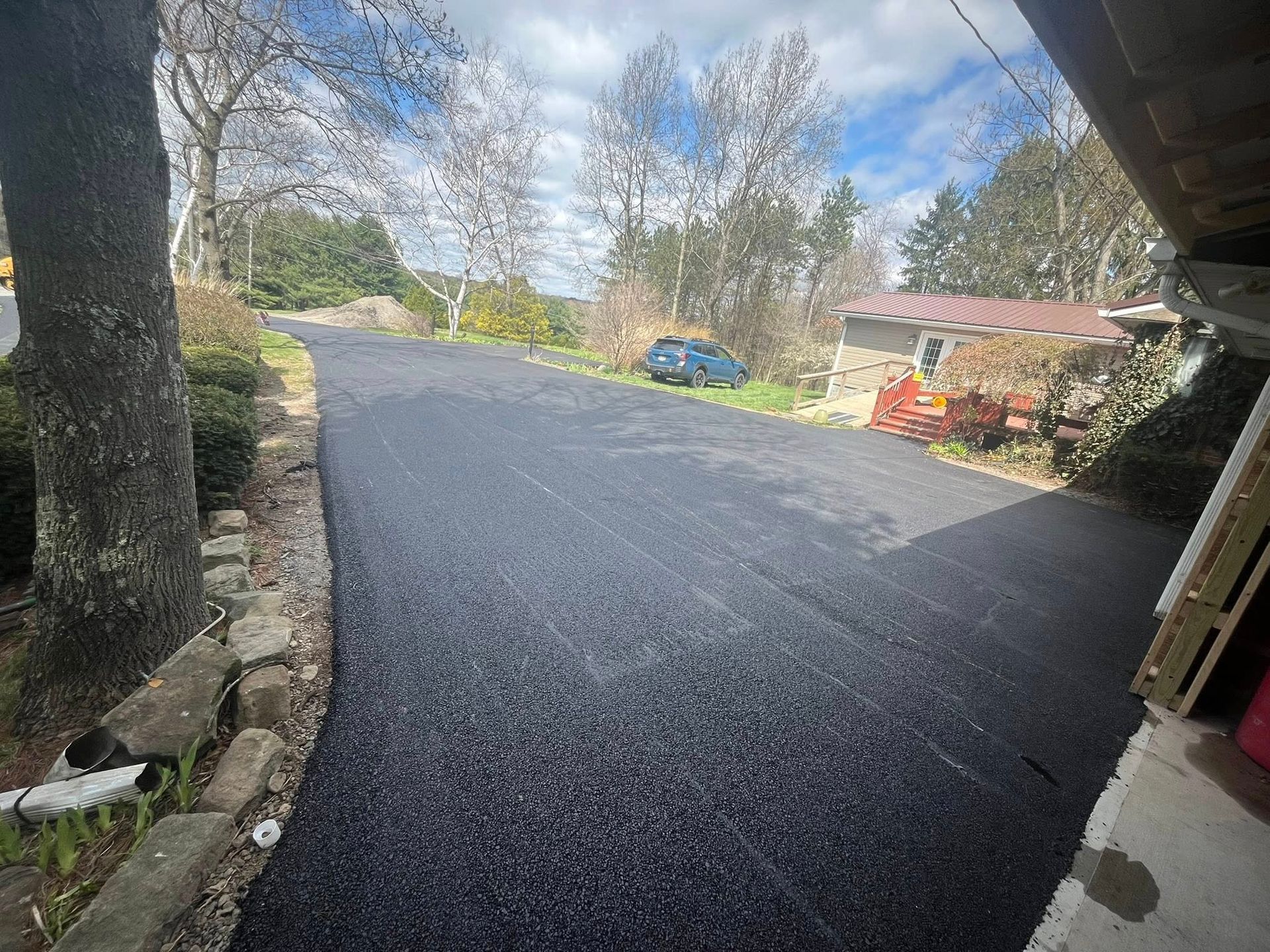 A newly paved asphalt driveway leads toward a house with a green car parked in the background under a blue sky.