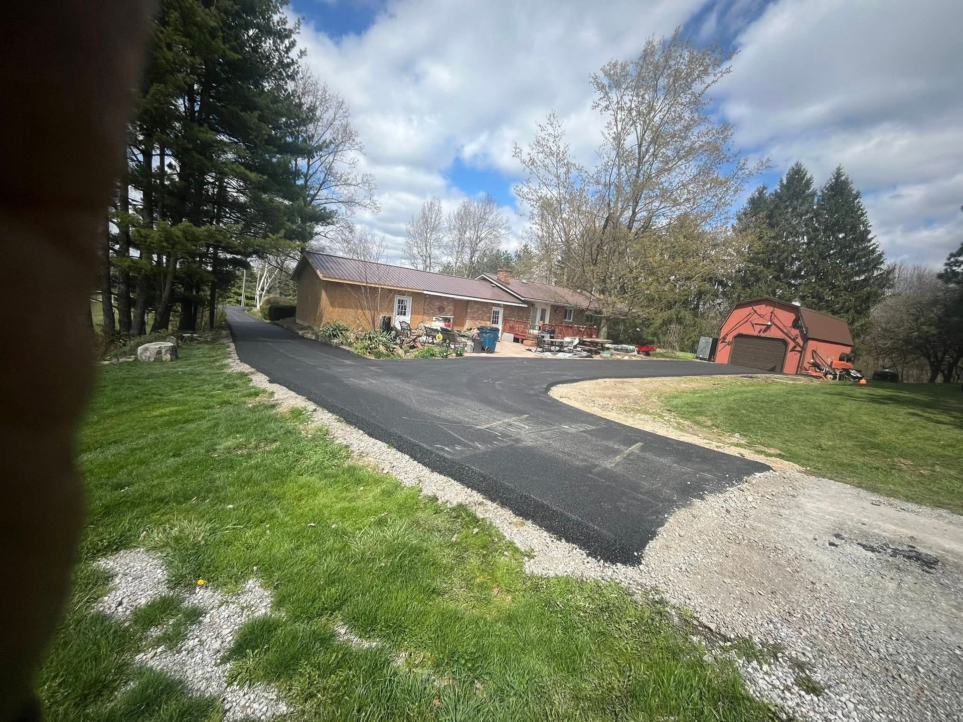 A freshly paved black asphalt driveway leads toward a one-story tan house and a small red barn on a grassy, wooded lot.