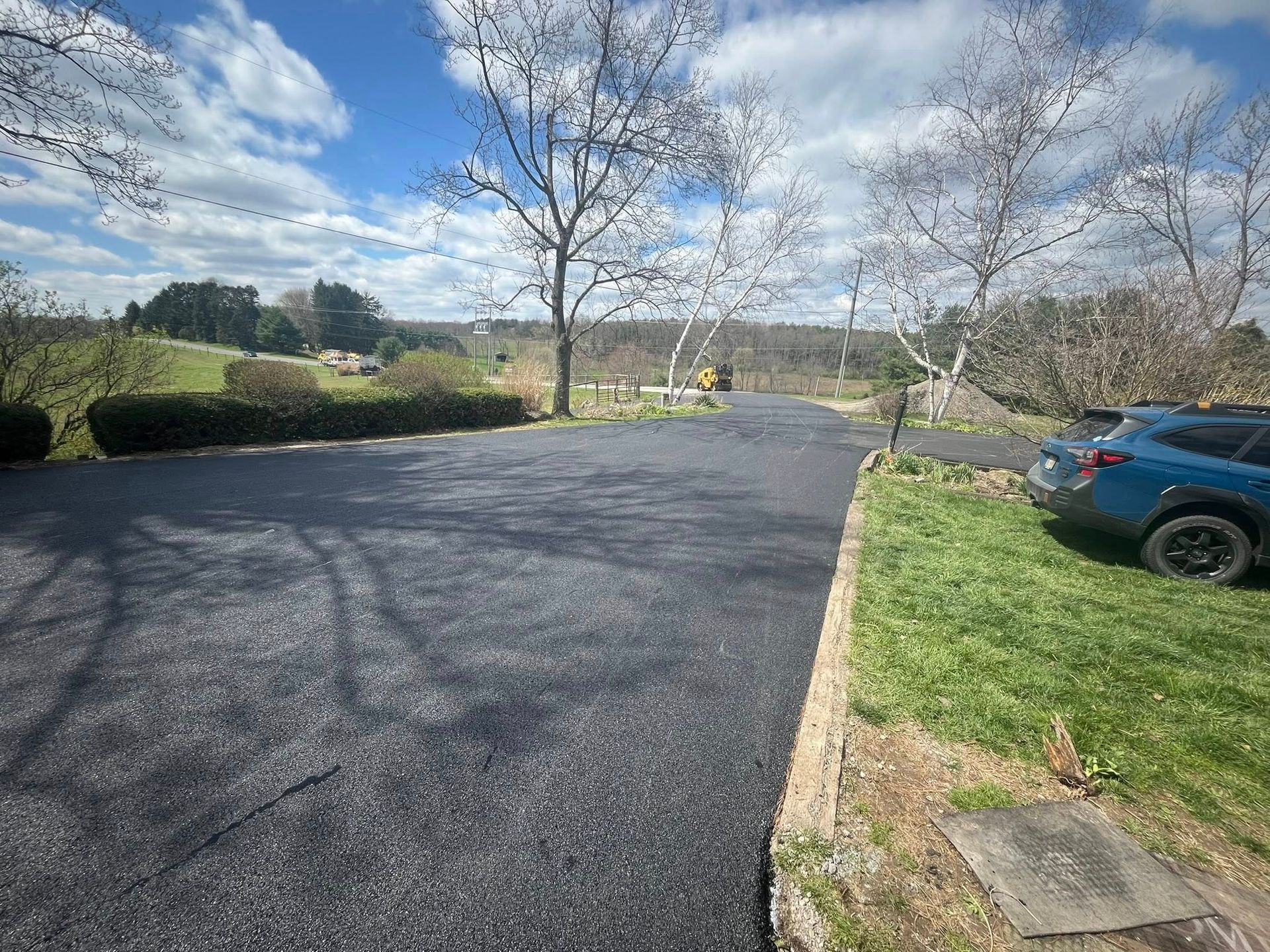 A newly paved asphalt driveway leads toward trees under a bright blue sky, with a blue car parked on the right.