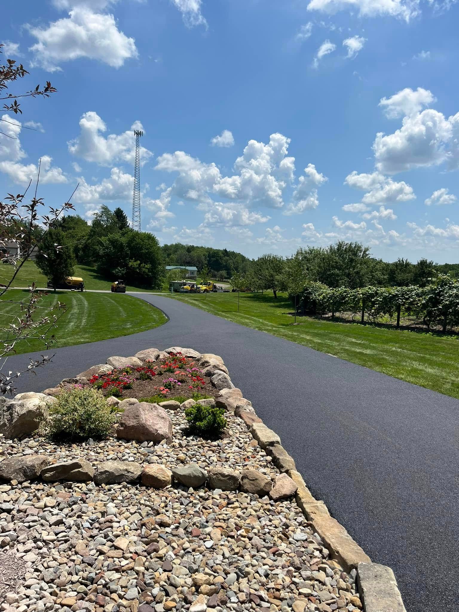An asphalt path leads through a landscape with a landscaped rock garden in the foreground, trees, and a blue, cloudy sky.