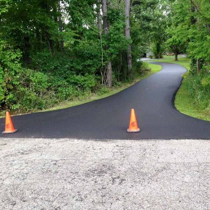 A newly paved, dark asphalt driveway leads into a tree-lined path, blocked by two orange traffic cones at the entrance.