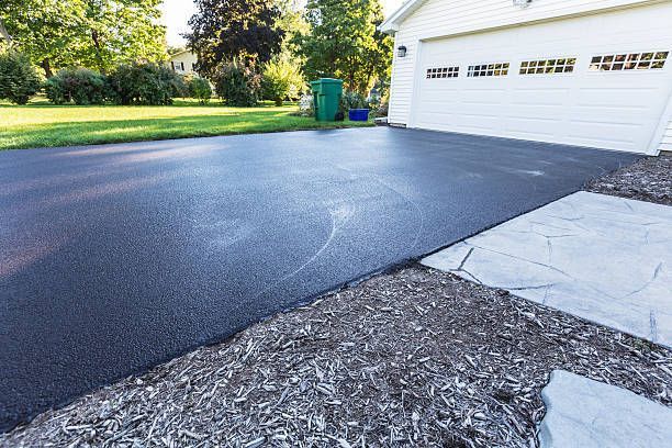 A freshly paved asphalt driveway leading to a white garage, viewed from a mulch-covered front yard.