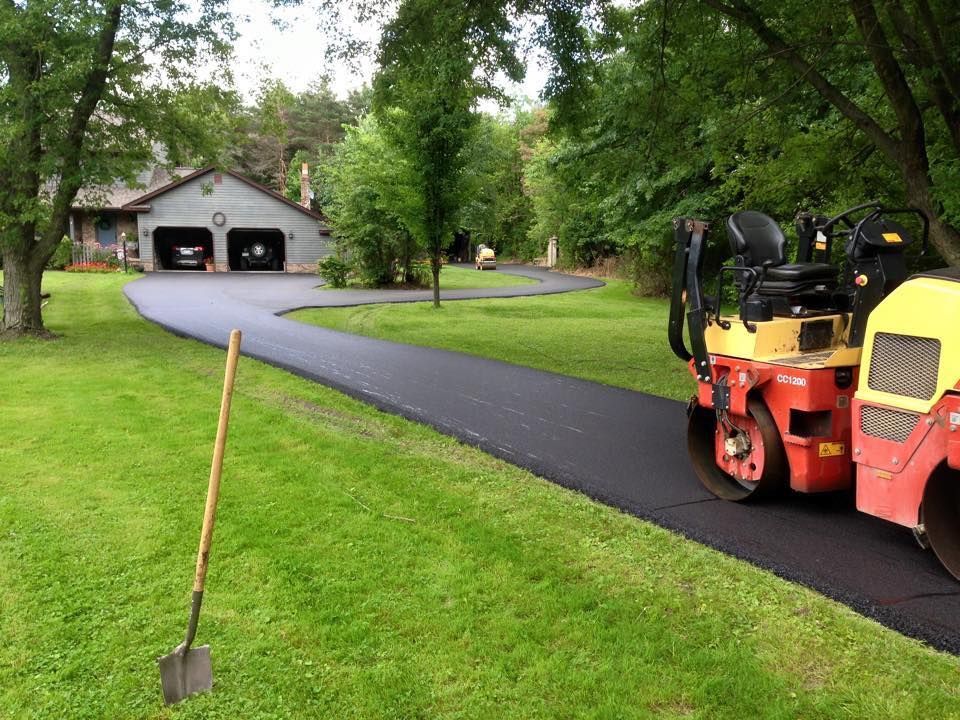 A yellow and red asphalt roller sits on a freshly paved driveway leading to a suburban home with a garage.