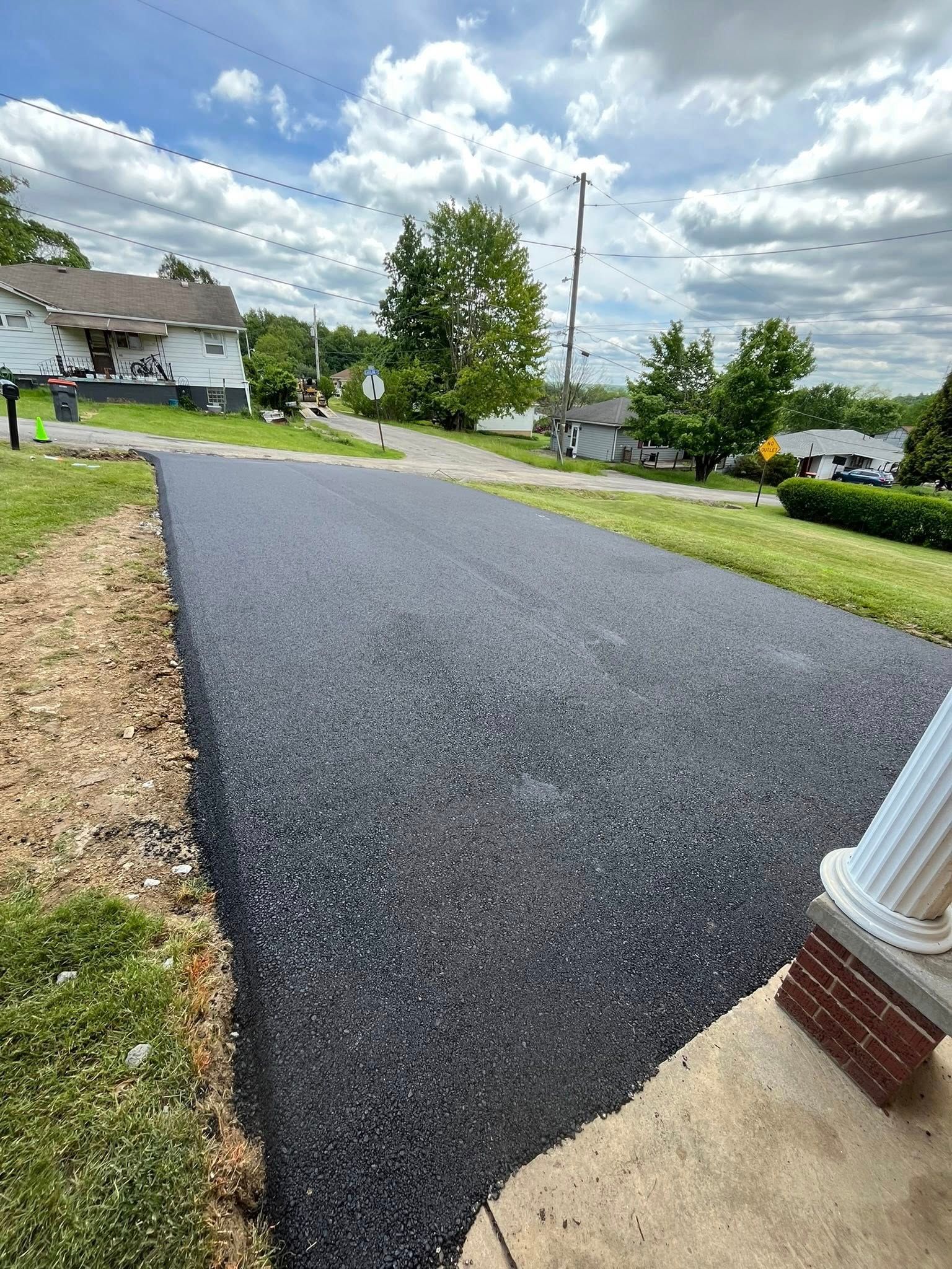 A newly paved black asphalt driveway leading up from a residential house lawn toward a road on a sunny, cloudy day.