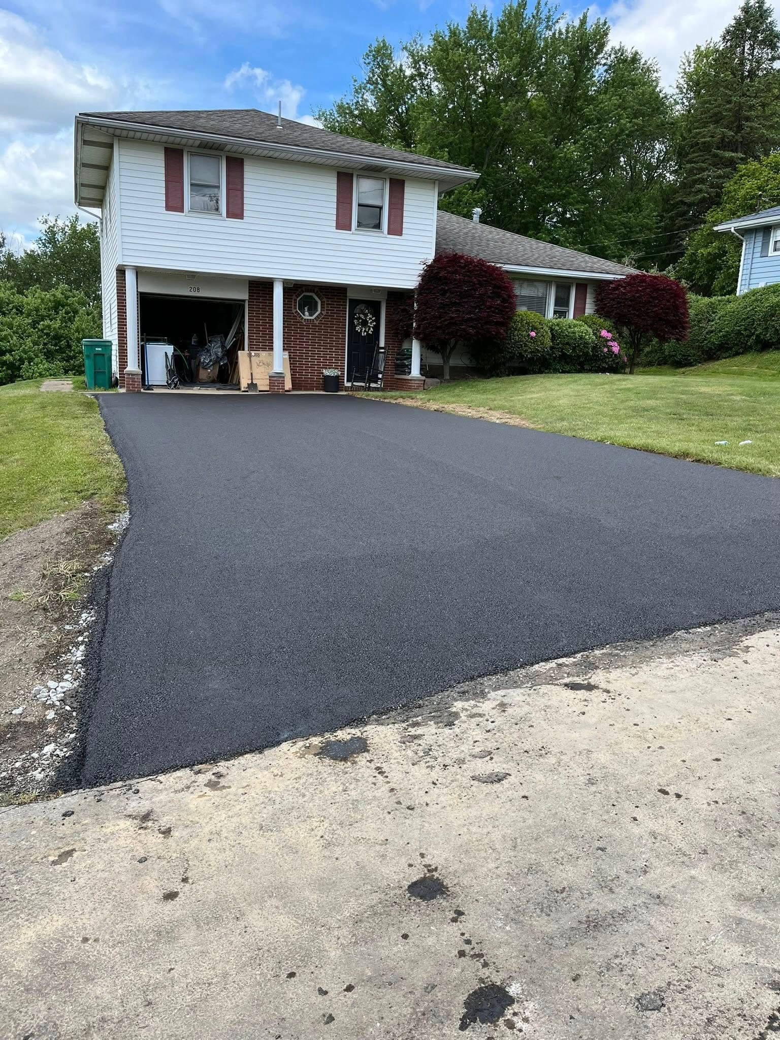 A newly paved asphalt driveway leads to the garage of a white two-story house on a sunny day with green landscaping.