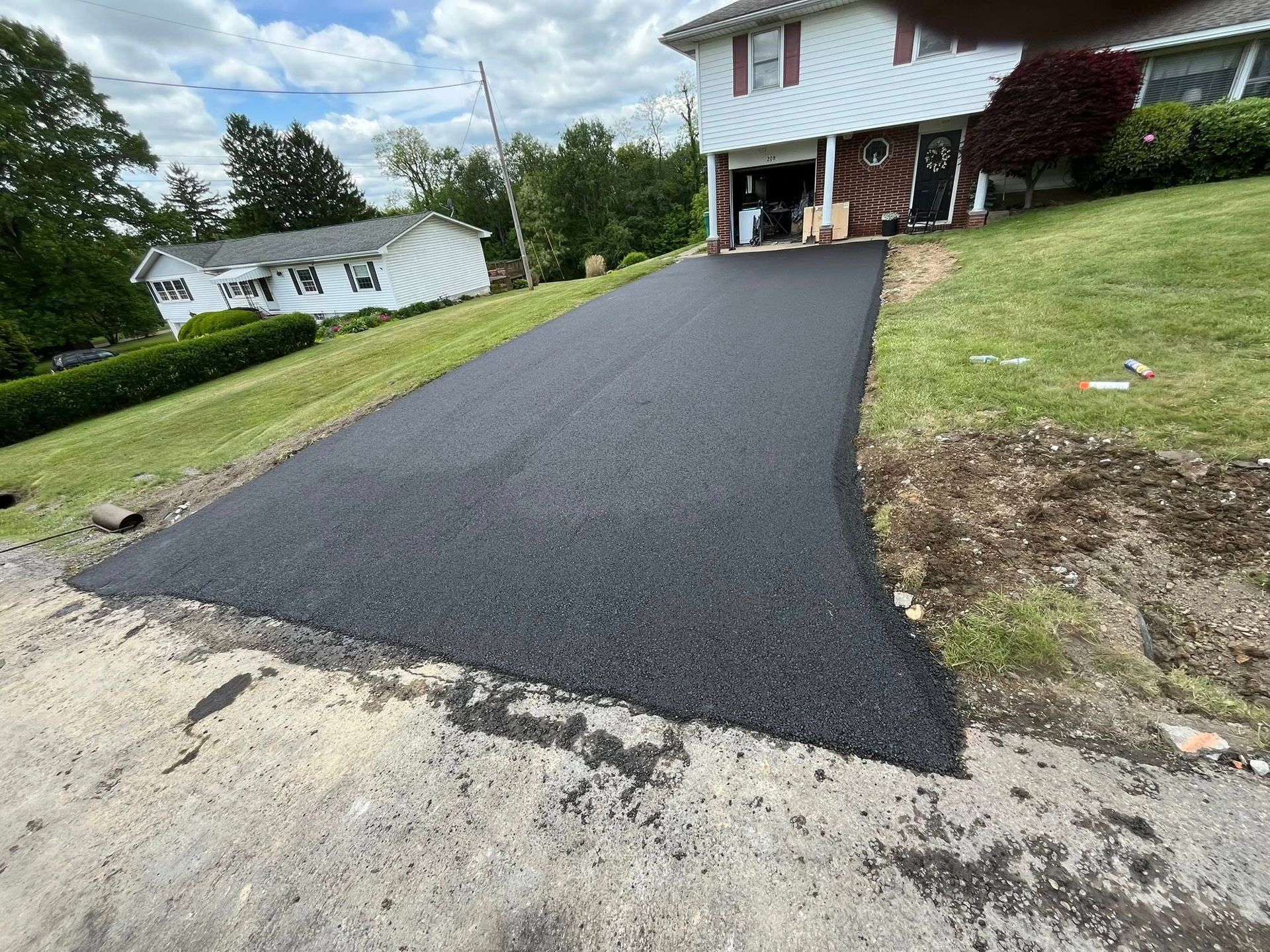A freshly paved, dark asphalt driveway leading up to a house on a sunny day with green lawns on both sides.