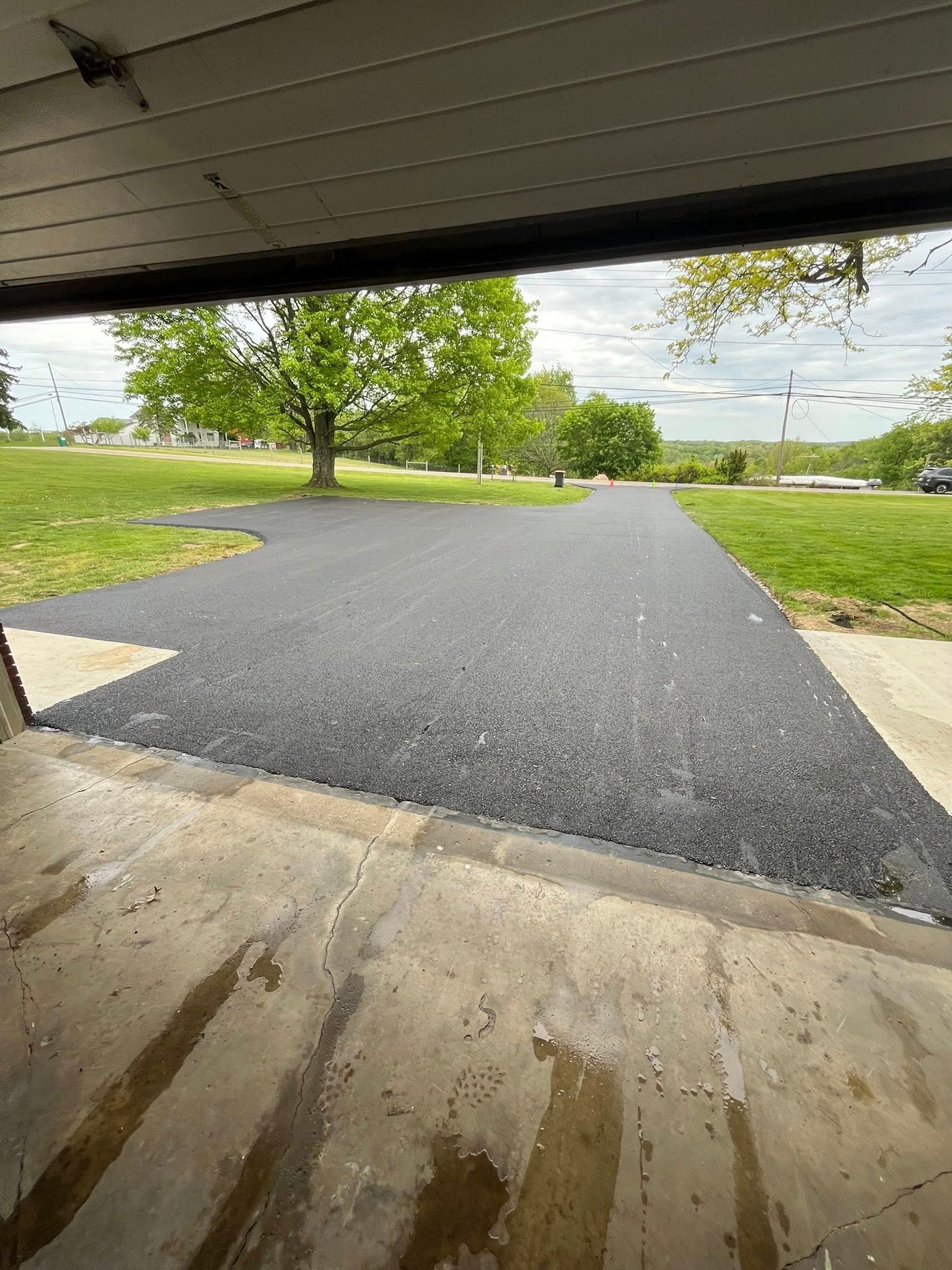 A newly paved asphalt driveway seen from a garage entrance, leading toward a grassy yard and trees under a cloudy sky.