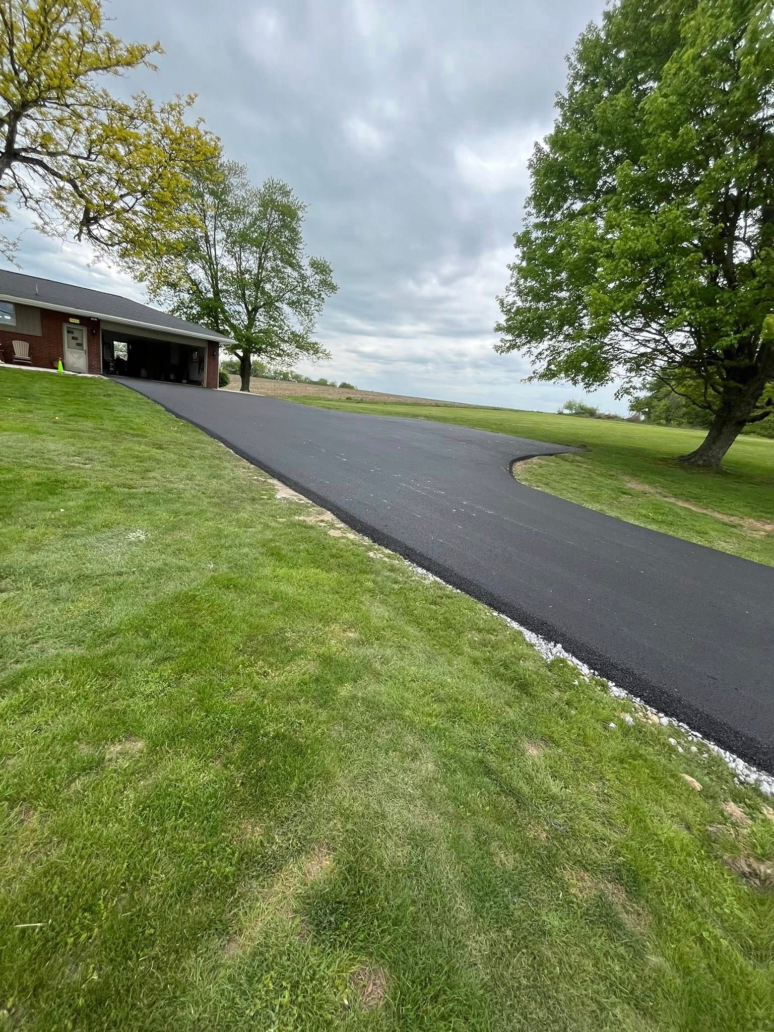 A freshly paved black asphalt driveway curves uphill towards a house, bordered by green grass and trees under a cloudy sky.