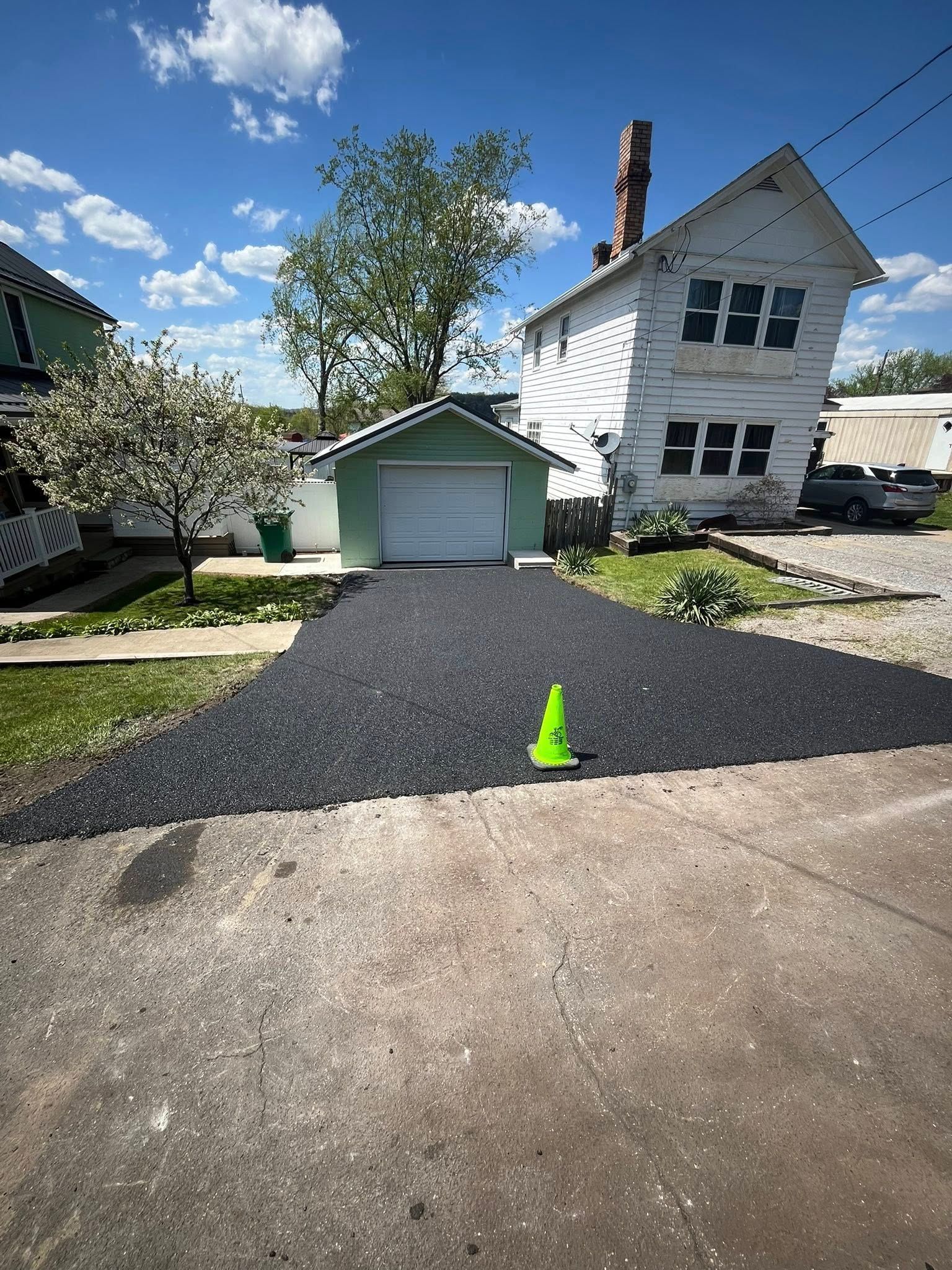A freshly paved, black driveway leads to a small green garage next to a white house under a blue, cloudy sky.
