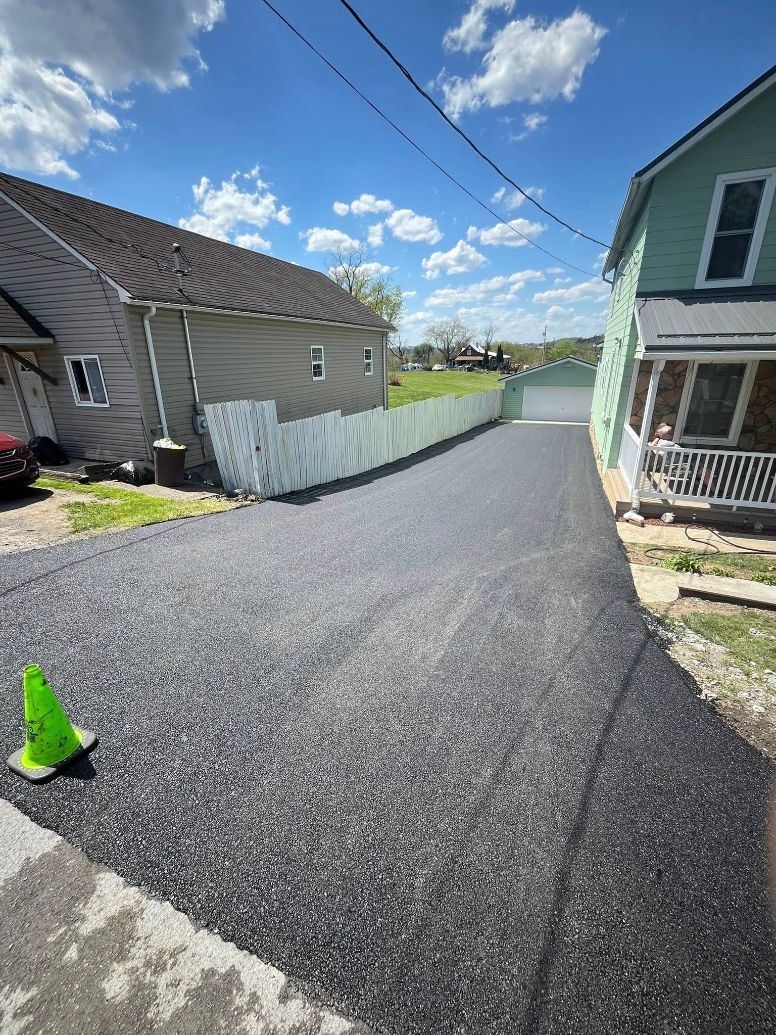 A newly paved asphalt driveway leads between two houses toward a white detached garage under a bright, cloudy sky.