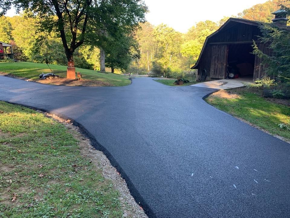 A newly paved asphalt driveway splits into two paths, leading toward a rural barn and trees under a clear, sunny sky.