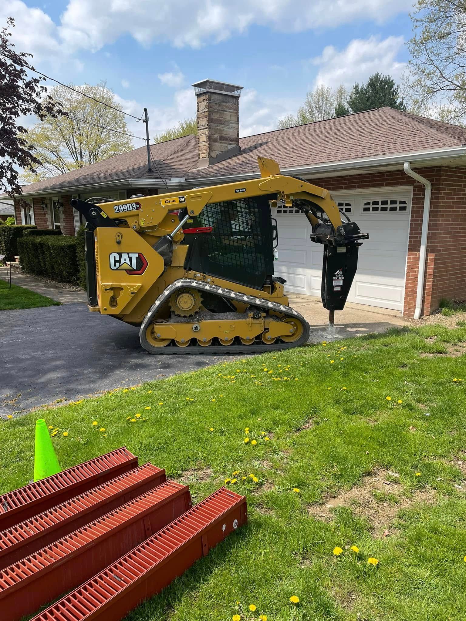 A yellow Caterpillar tracked skid steer with a hydraulic hammer attachment on a residential driveway near a brick home.