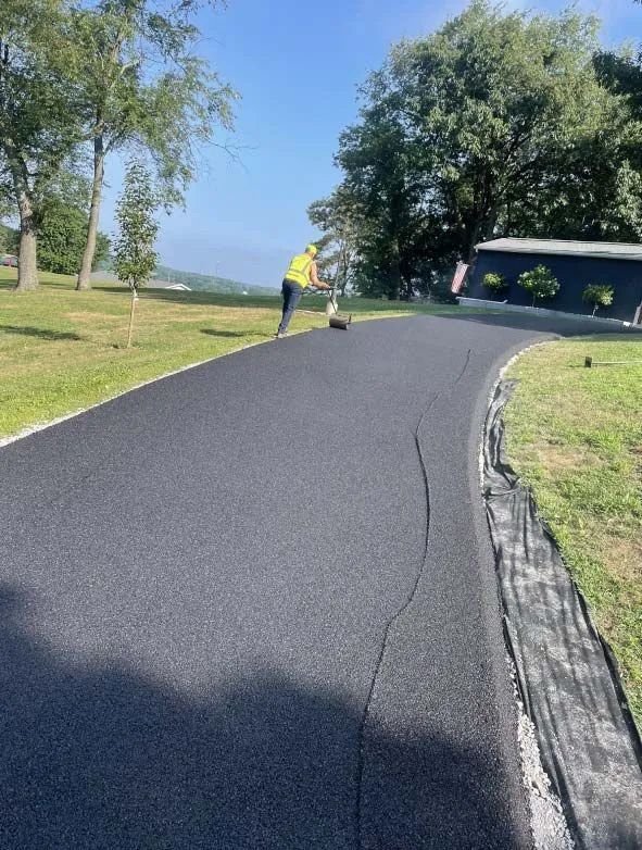 A worker in a high-visibility vest uses a walk-behind roller on a freshly paved, dark asphalt driveway.