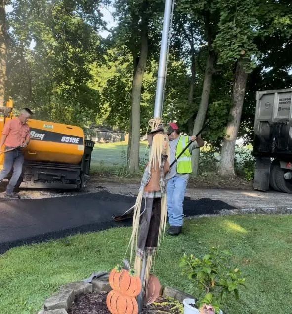 Two workers in construction gear operate a yellow paving machine on a driveway, with a decorative scarecrow in the foreground.