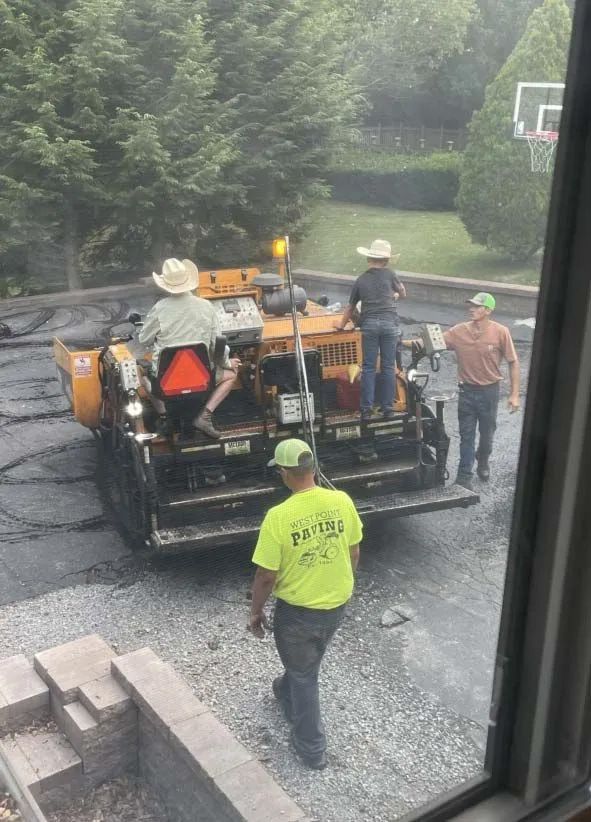 Three workers in construction gear operate a yellow paving machine on a gravel driveway near a brick retaining wall.