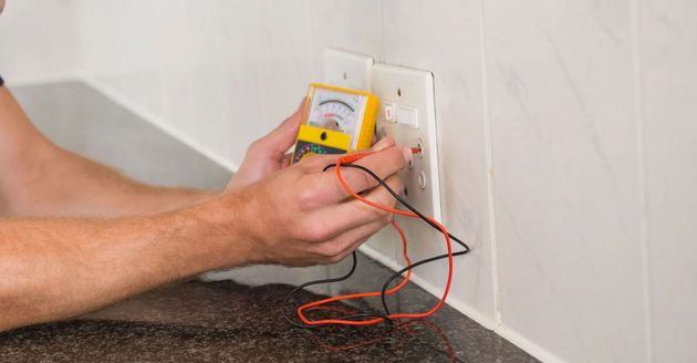 Person testing an electrical outlet with a multimeter and red/black leads near a wall.