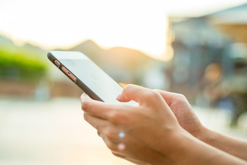 Hands holding and using a smartphone with a blurry outdoor background and golden sunlight.