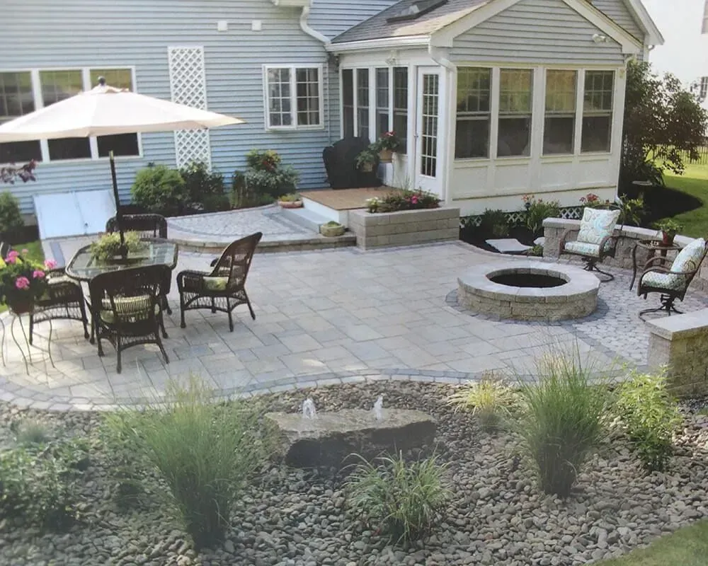 Backyard patio with fire pit, seating, water feature, and house in background.