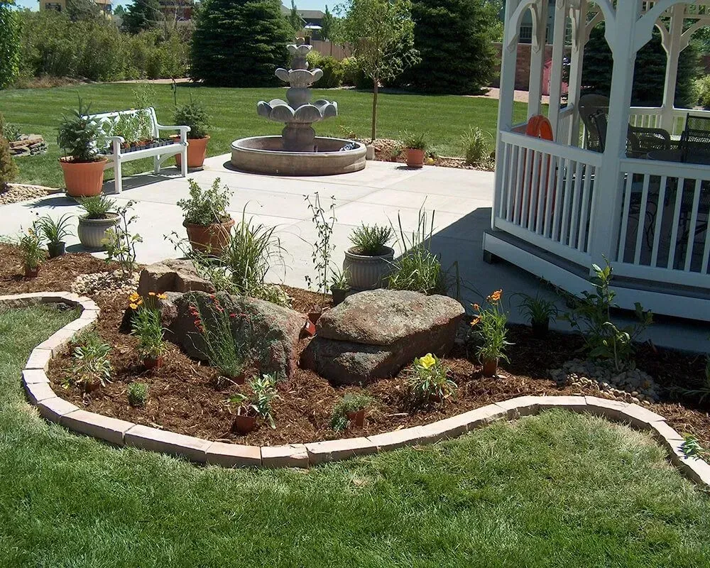 A backyard with a flower bed, fountain, gazebo, and green lawn.