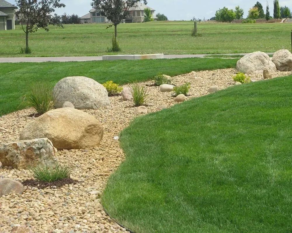 Lawn with a curved edge bordered by rocks and gravel, with a grassy background and distant houses.