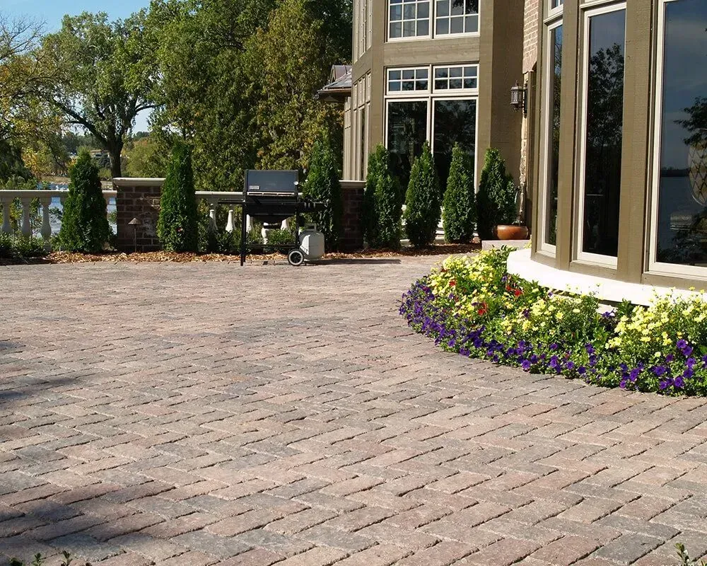 Brick patio with a grill and flower bed in front of a house with large windows.