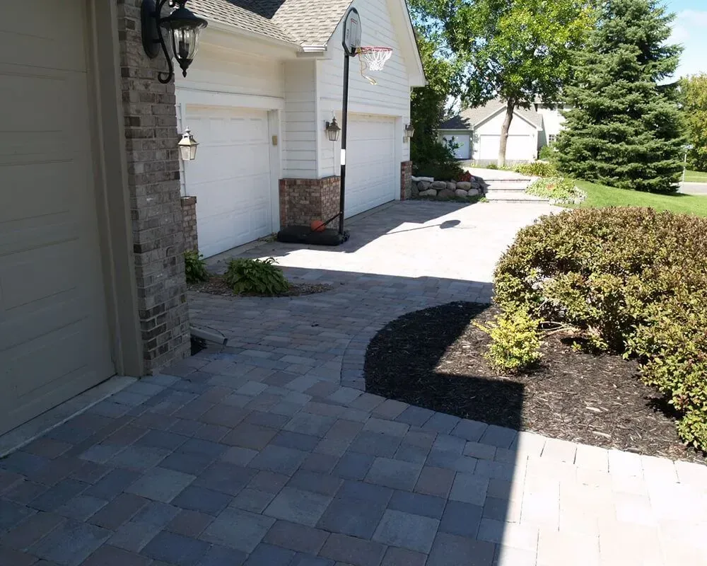 Brick paver walkway leading to a house with a basketball hoop and garage doors, surrounded by landscaping.