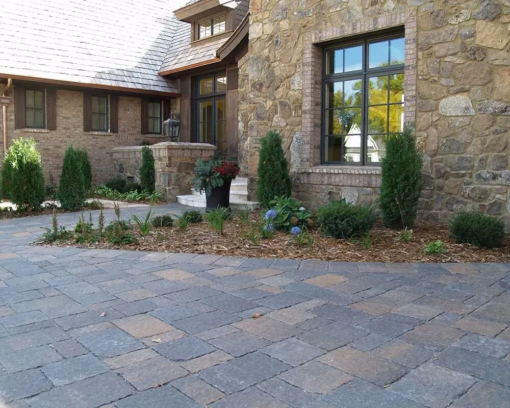 Stone house with a paved driveway and landscaped yard with shrubs and a large window.
