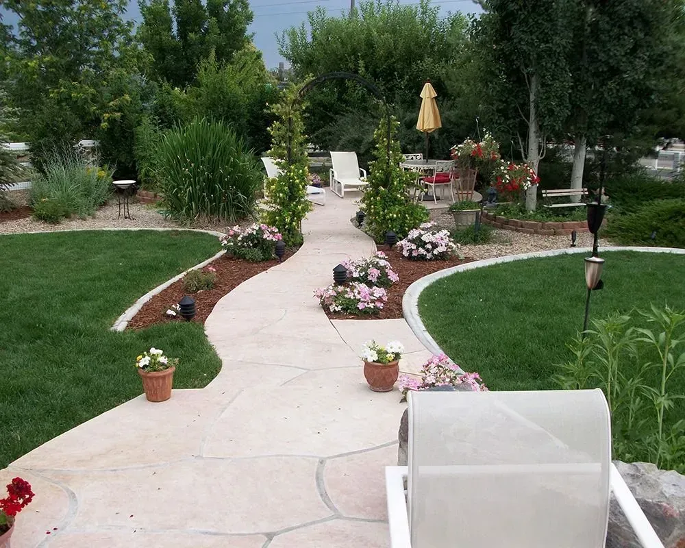 Stone pathway winds through a lush backyard with flowerbeds, green grass, and outdoor seating.