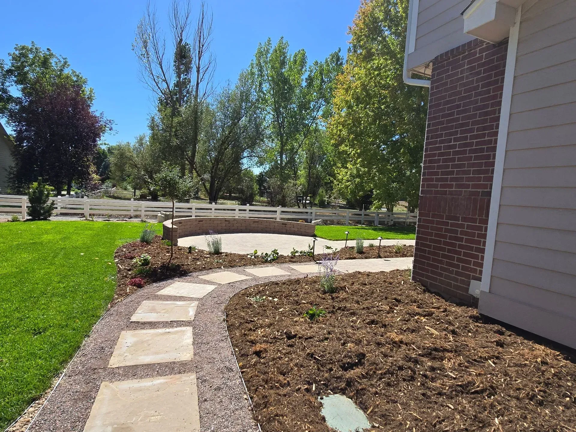 Stone pathway leads to a circular patio bordered by a landscaped yard with green grass and trees.