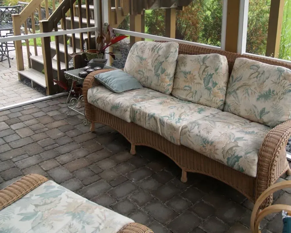 Wicker sofa with floral cushions on a screened porch. Brick flooring and wooden stairs visible.