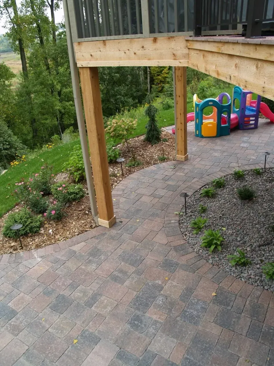 Brick pathway curves around wooden deck supports, leading toward a playground.