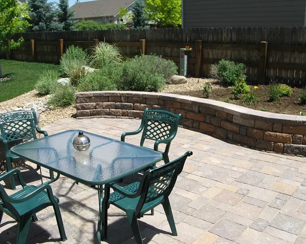 Patio with glass table, green chairs, brick retaining wall, and landscaped garden.