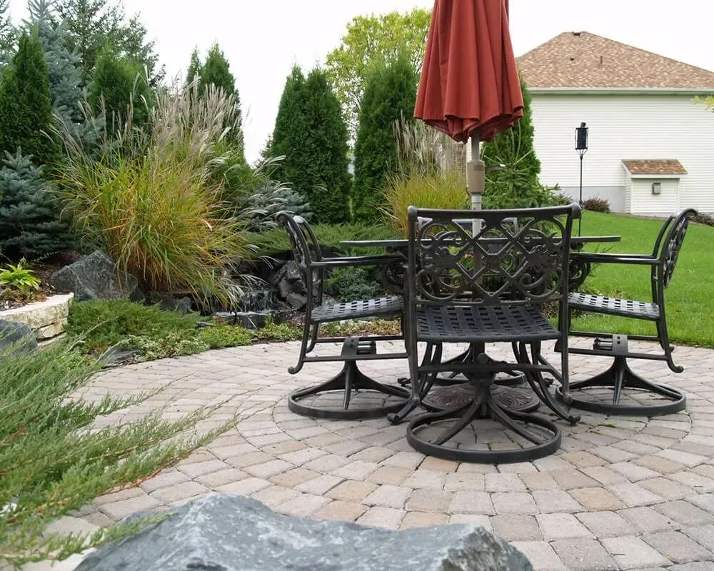 Patio set on circular stone patio with umbrella, plants, and small waterfall.