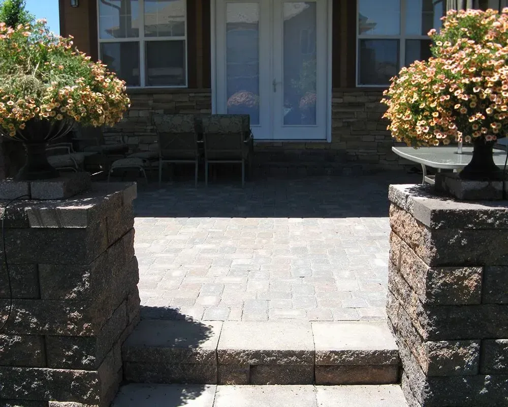 Stone patio with steps, bordered by brick pillars holding flowering plants; back doors visible.