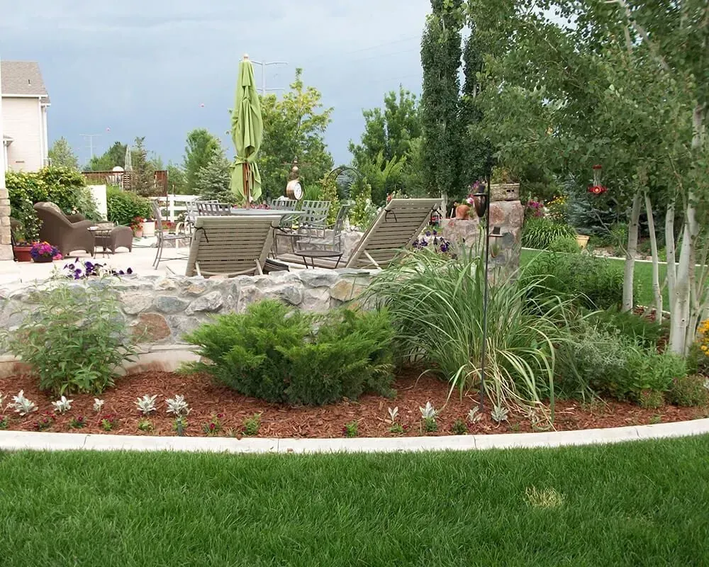 Backyard garden with green grass, rock wall, patio furniture, and trees under a cloudy sky.