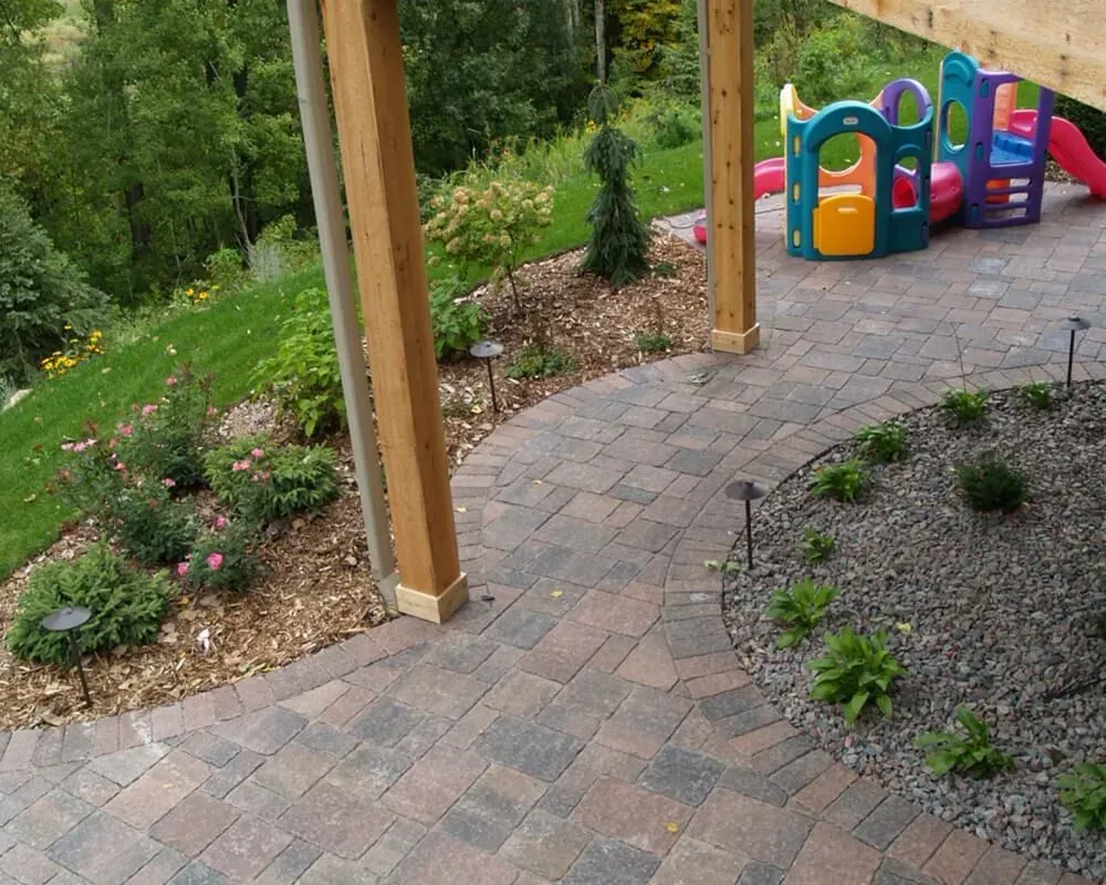 Brick patio with landscaping and playground equipment in the background. Wooden posts with light fixtures.