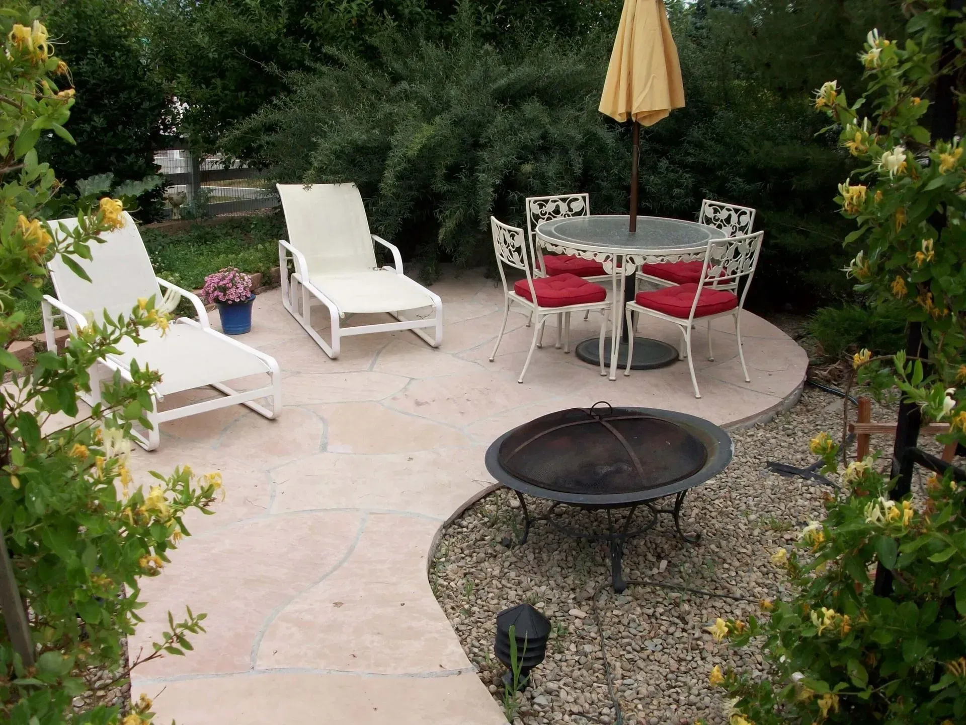 Patio with lounge chairs, round table, fire pit, and umbrella surrounded by landscaping.