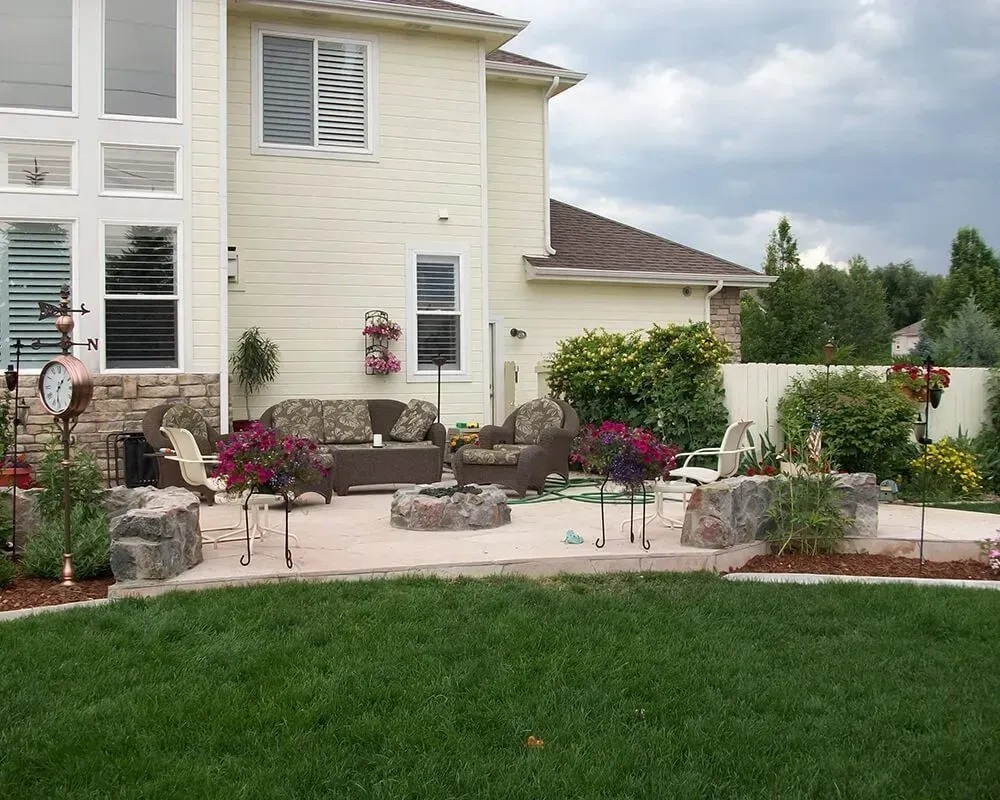Patio with outdoor furniture, fire pit, and hanging flower baskets; beige house in background.