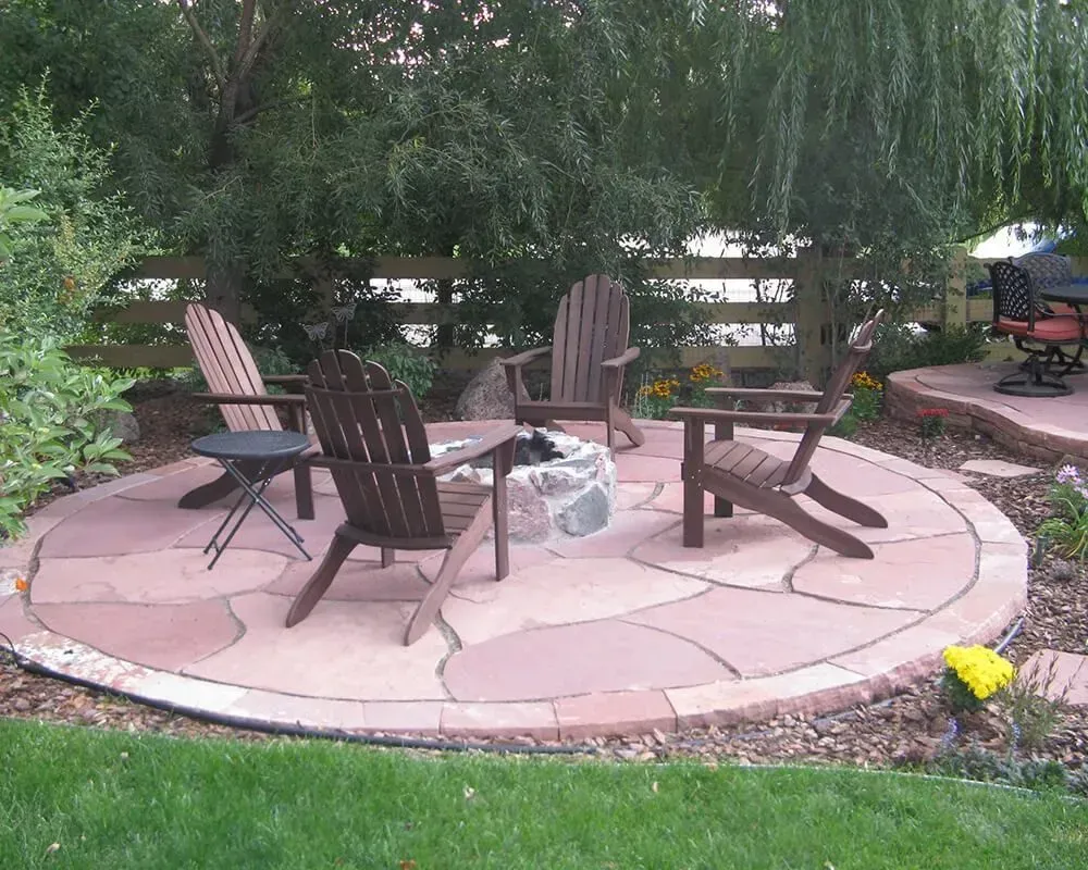 Round stone patio with Adirondack chairs around a fire pit, in a backyard setting.