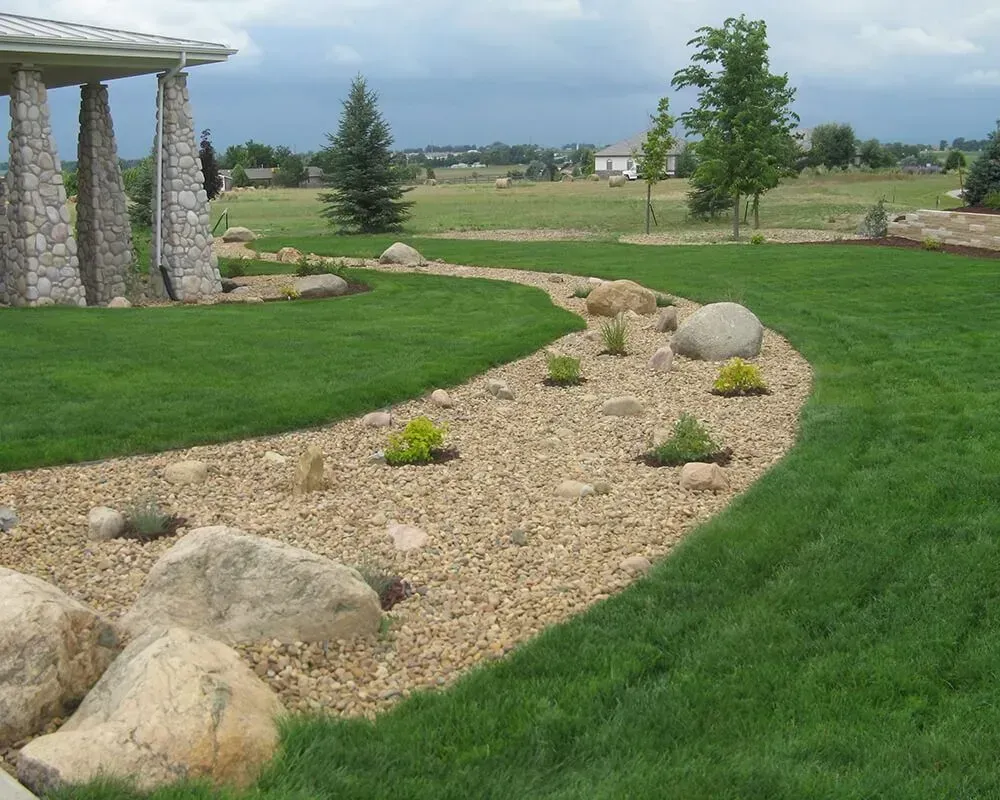 Rock garden with tan gravel, large boulders, and small green plants; green lawn and trees in the background.