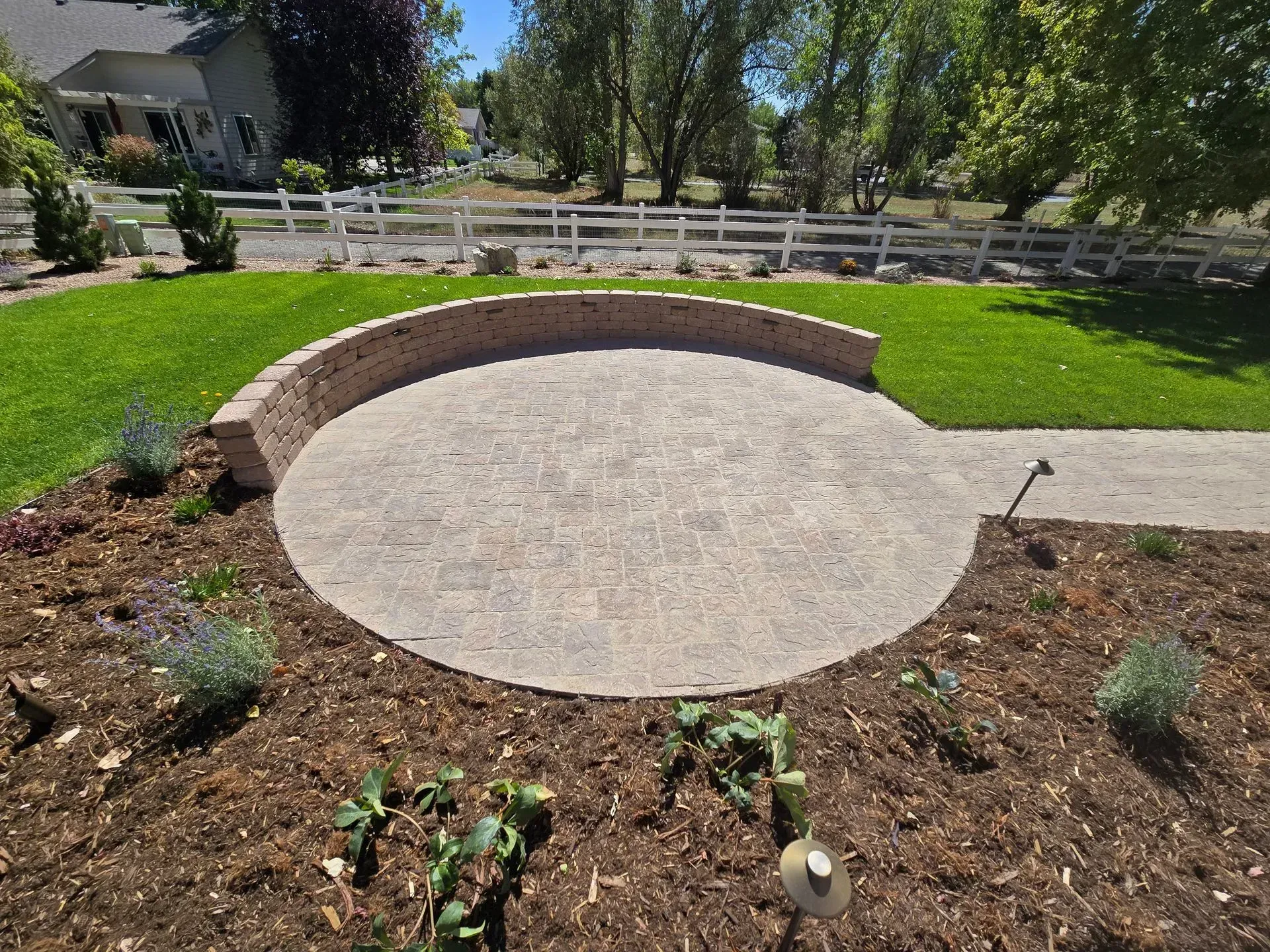 Circular brick patio with retaining wall, surrounded by grass and flower beds.