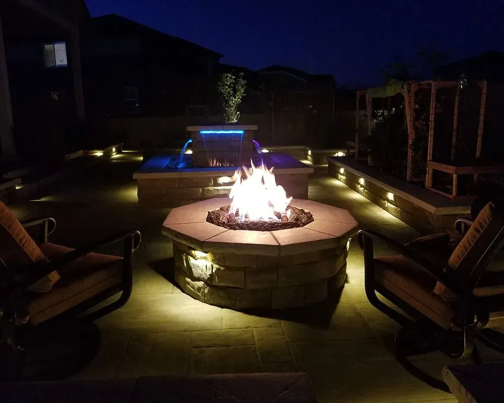 Nighttime backyard scene with lit fire pit, water fountain, and outdoor seating.