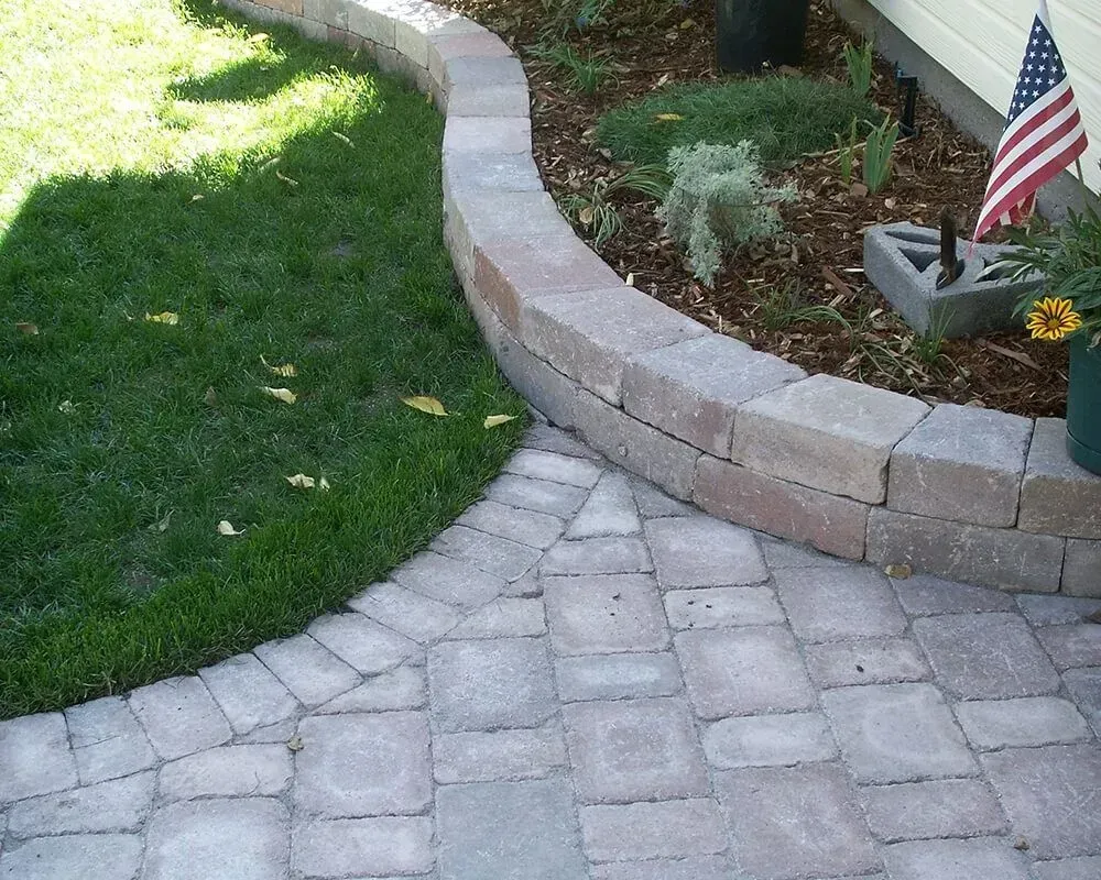 Curved brick retaining wall bordering grass and a paved path, with a garden bed and American flag.