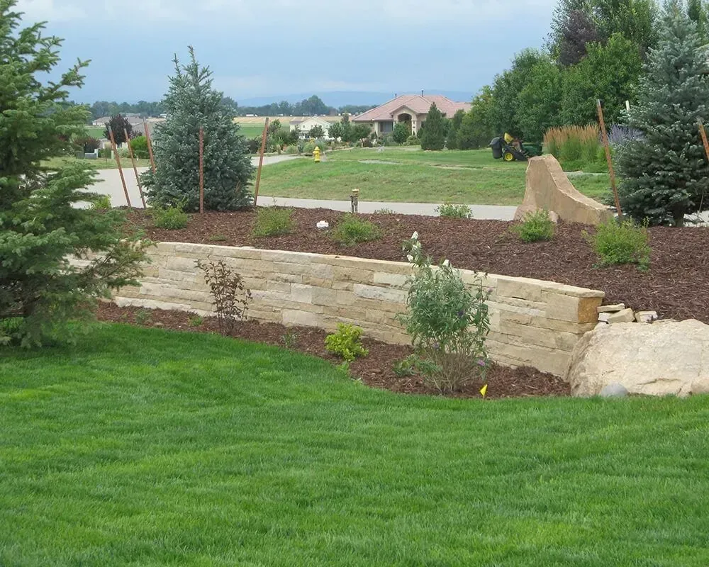 Stone retaining wall in a front yard with trees and grass, residential setting.