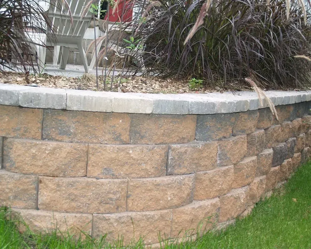 Curved retaining wall of brown blocks topped with gray capstones, supporting a garden bed.