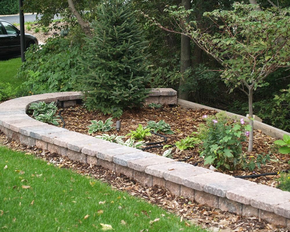 Curved brick retaining wall with garden bed, green grass, and trees.